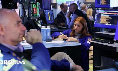 In the left corner, a bald male trader, and a woman with auburn hair, also a trader, in the middle, holding a pen and writing into a notebook, on the floor of the New York Stock Exchange.