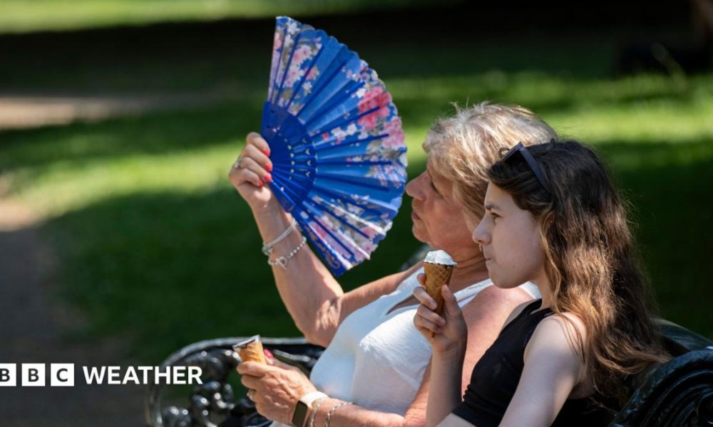 two women sitting on a park bench.  One eating an ice cream, the other fanning herself in the hot weather