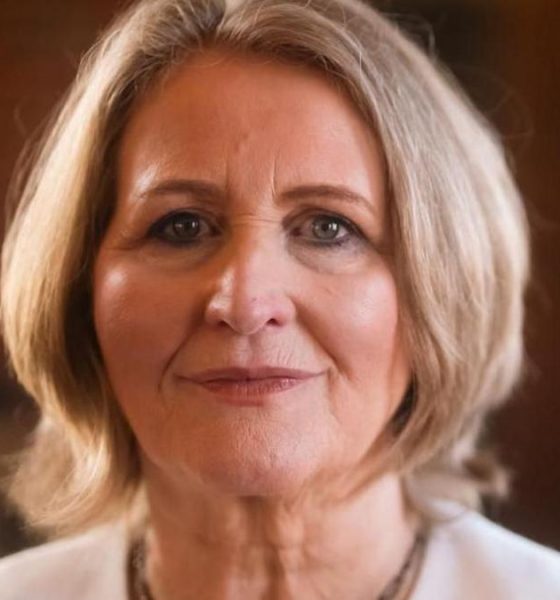 A woman with short blonde hair wearing a white suit jacket smiles faintly at the camera