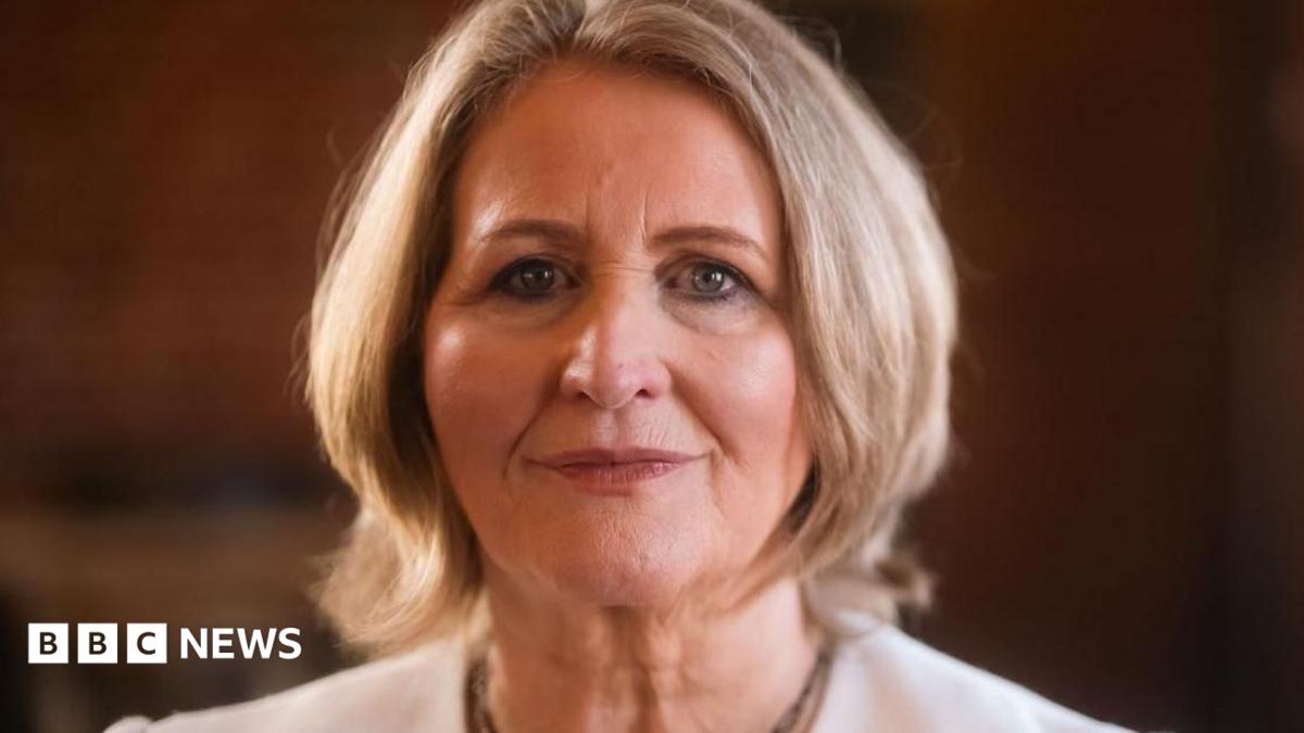 A woman with short blonde hair wearing a white suit jacket smiles faintly at the camera