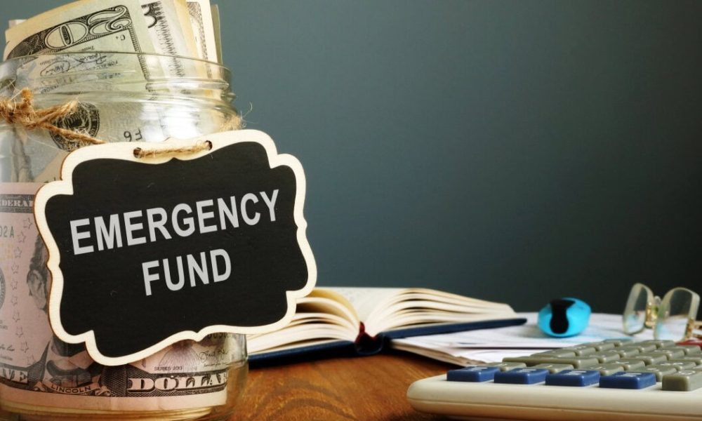glass jar filled with money has a sign reading "emergency fund;" in the background a book is open and glasses are on a table near a calculator.