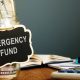 glass jar filled with money has a sign reading "emergency fund;" in the background a book is open and glasses are on a table near a calculator.