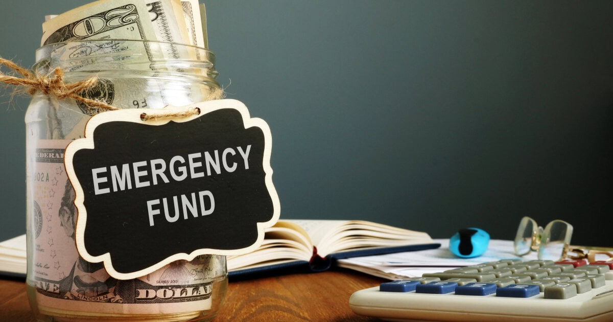 glass jar filled with money has a sign reading "emergency fund;" in the background a book is open and glasses are on a table near a calculator.