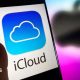 A phone with the words iCloud in black letter, and the iCloud blue and white logo of a cloud on it, against a white background. A blurred Apple logo in black is behind it against a purple background.