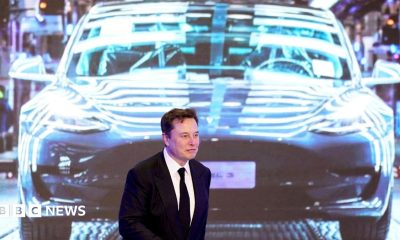 Elon Musk walking in front of a projected image of a Tesla car, wearing a black suit and tie.