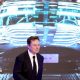Elon Musk walking in front of a projected image of a Tesla car, wearing a black suit and tie.