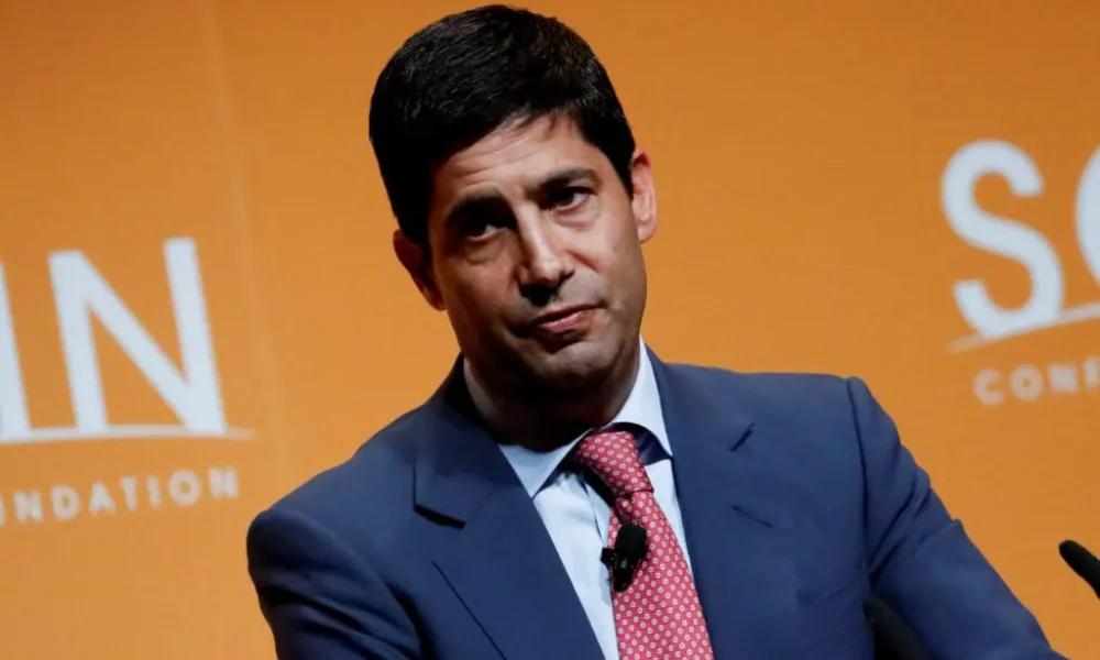 A man in a suit speaking at an event with an orange background.