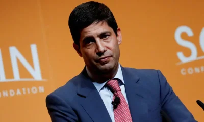 A man in a suit speaking at an event with an orange background.