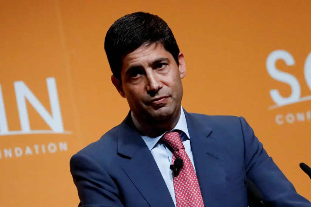 A man in a suit speaking at an event with an orange background.