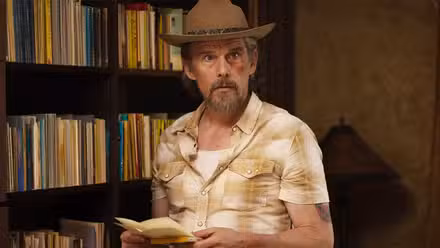 Lee Raybon (Ethan Hawke) with a bruised face and a hat, standing in front of bookshelves in 'The Lowdown'