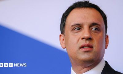 Anas Sawar stares into the distance. He has dark hair and is wearing a dark suit and white shirt. The background is blue and white.
