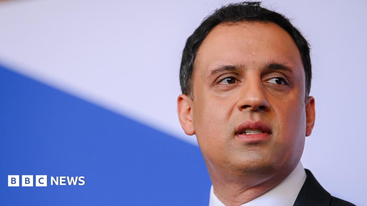 Anas Sawar stares into the distance. He has dark hair and is wearing a dark suit and white shirt. The background is blue and white.