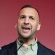 A woman with short brown hair looks directly into the camera with a slight smiling expression. She is wearing a pink top and a silver necklace with a heart charm.