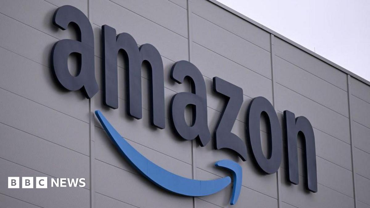 The Amazon logo, which is the world 'Amazon' with its signature stylised arrow underneath, on the side of a grey building with a light overcast sky in the background