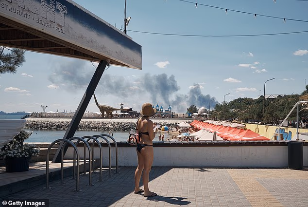 Local residents and visitors relax on a beach on the outskirts of Odesa as smoke from a fire rises in the background following a Russian missile strike during an air raid alert, on July 29, 2026 in Odesa, Ukraine
