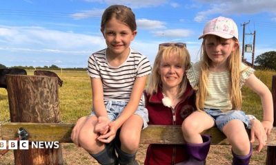 Heather Oldfield stands in the centre of the image wearing a burgundy fleece gilet and sunglasses on her head. On either side of her is a young child. They are in a field with cattle behind them.