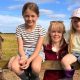 Heather Oldfield stands in the centre of the image wearing a burgundy fleece gilet and sunglasses on her head. On either side of her is a young child. They are in a field with cattle behind them.