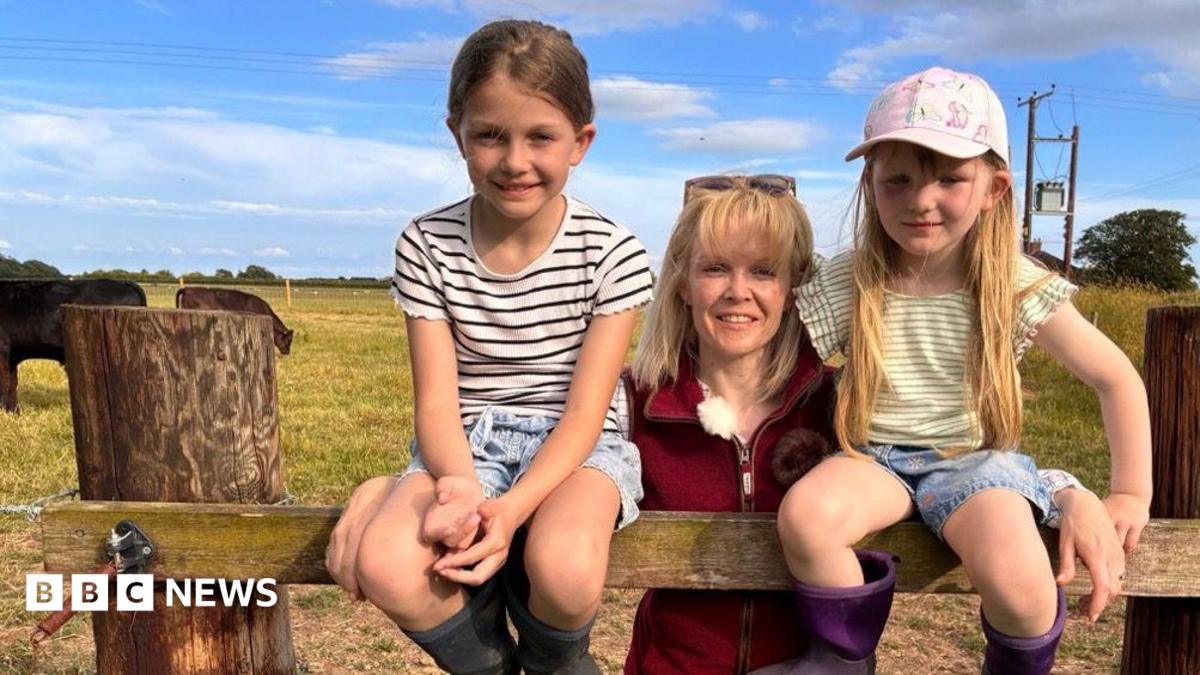 Heather Oldfield stands in the centre of the image wearing a burgundy fleece gilet and sunglasses on her head. On either side of her is a young child. They are in a field with cattle behind them.