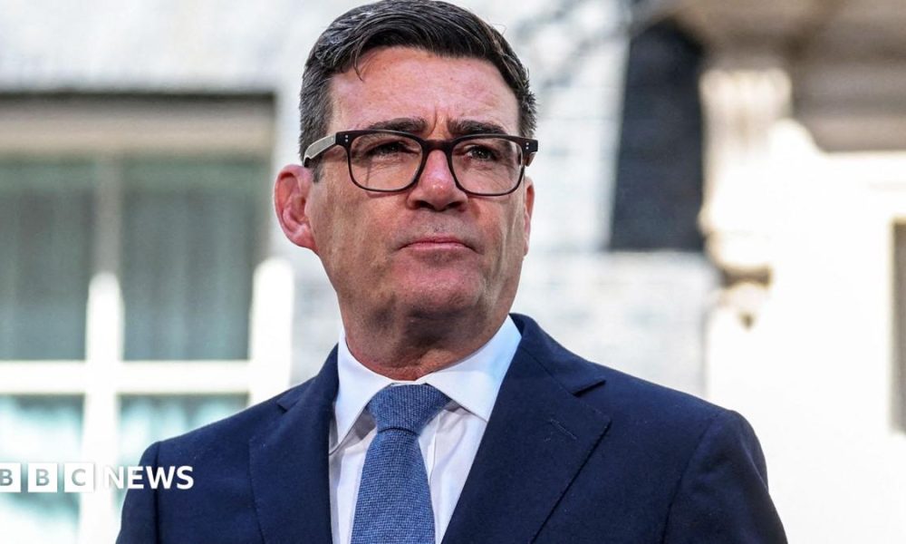Prime Minister Andy Burnham wears dark-rimmed glasses and a blue suit.