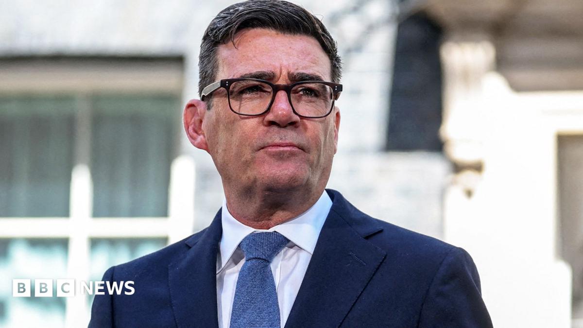Prime Minister Andy Burnham wears dark-rimmed glasses and a blue suit.