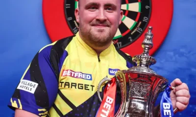 Luke "the Nuke" Littler smiles with his winner's trophy in hand infront of a darts board in Blackpool