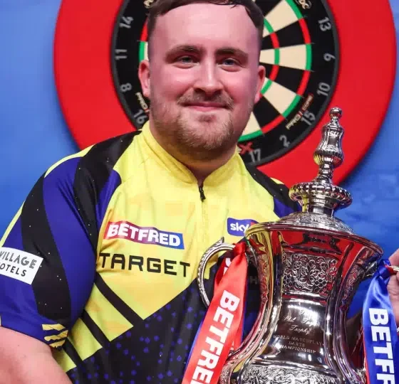 Luke "the Nuke" Littler smiles with his winner's trophy in hand infront of a darts board in Blackpool