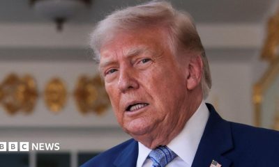 US President Donald Trump speaking in front of a White House entrance, wearing a blue suit and tie.