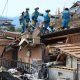 Rescue workers in overalls stand on top of a collapsed house