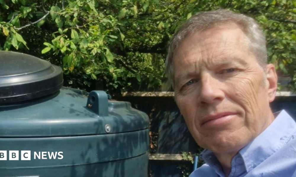 A man in a blue shirt takes a selfie in a garden next to a green oil tank. He has grey hair and there are leaves behind the tank.