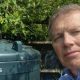 A man in a blue shirt takes a selfie in a garden next to a green oil tank. He has grey hair and there are leaves behind the tank.