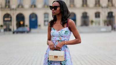 PARIS, FRANCE - APRIL 29: Emilie Joseph wears black large sunglasses, crystal earrings, a pale blue with red / green / navy blue / beige flower print pattern heart neck / tank-top / belted / midi dress, a beige wickers and white shiny leather bi-material handbag, during a street style fashion photo session, on April 29, 2023 in Paris, France. (Photo by Edward Berthelot/Getty Images)