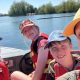 A woman wearing a yellow cap and three children, on a boat. All are wearing life jackets, and the photo is taken as a selfie-style with everyone looking at the camera