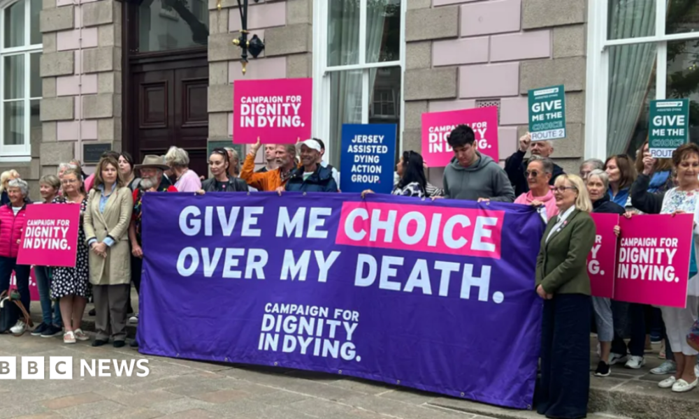 Campaigners for assisted dying holding a large purple "Give me choice over my death" and placards outside a grey building, the States Chamber.