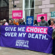 Campaigners for assisted dying holding a large purple "Give me choice over my death" and placards outside a grey building, the States Chamber.