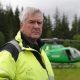 Nethy Bridge community council leader Sandy McCook wearing a high-visibility yellow jacket stands in a grassy clearing with a green helicopter parked behind them. Dense conifer forest forms the background under an overcast sky.