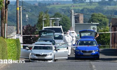A wide shot showing the Old Cullybackey Road with police tape across it. Three silver cars and a white van are parked one behind the other on the left of the road while a blue Skoda is parked in the middle of the road. Two forensic officers in white forensic suits can be seen as well as a PSNI officer.