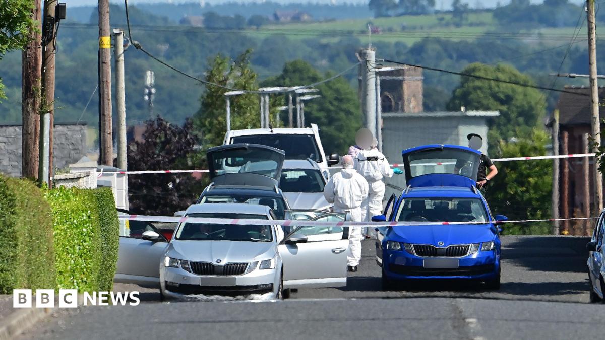 A wide shot showing the Old Cullybackey Road with police tape across it. Three silver cars and a white van are parked one behind the other on the left of the road while a blue Skoda is parked in the middle of the road. Two forensic officers in white forensic suits can be seen as well as a PSNI officer.