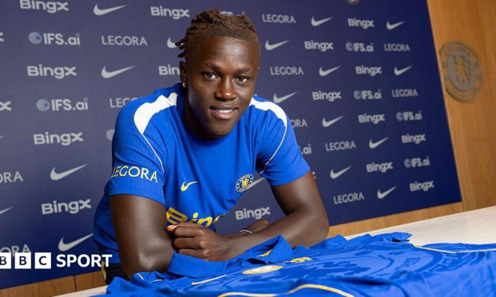 Quenda resting his arms on a table with a Chelsea shirt on and in front of him after signing for the club