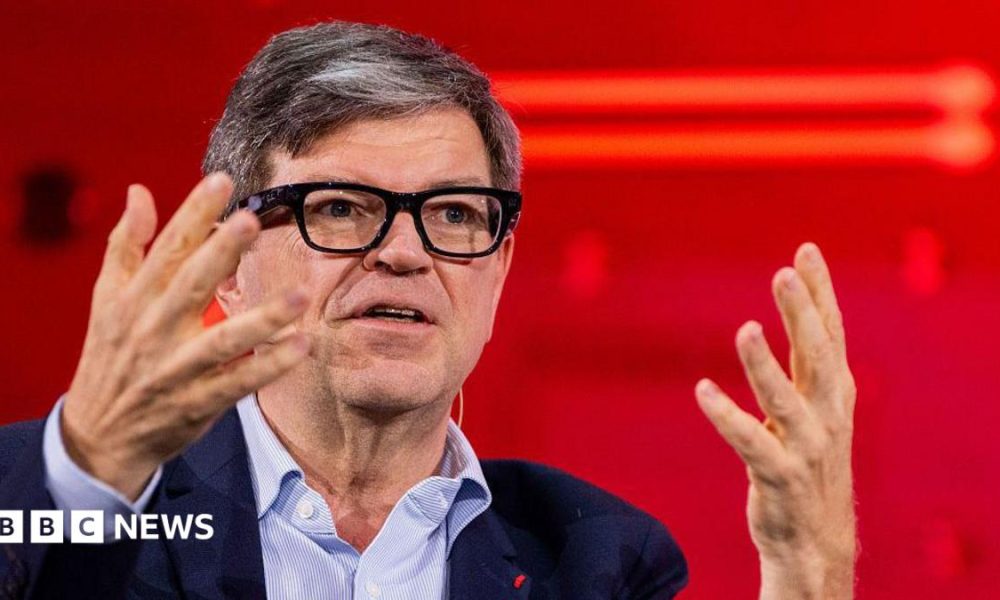 Yann LeCun wears fashionable thick-rimmed glasses, a light blue shirt and navy jacket. He is using his hands to articulate a point while speaking at a conference.