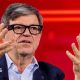Yann LeCun wears fashionable thick-rimmed glasses, a light blue shirt and navy jacket. He is using his hands to articulate a point while speaking at a conference.