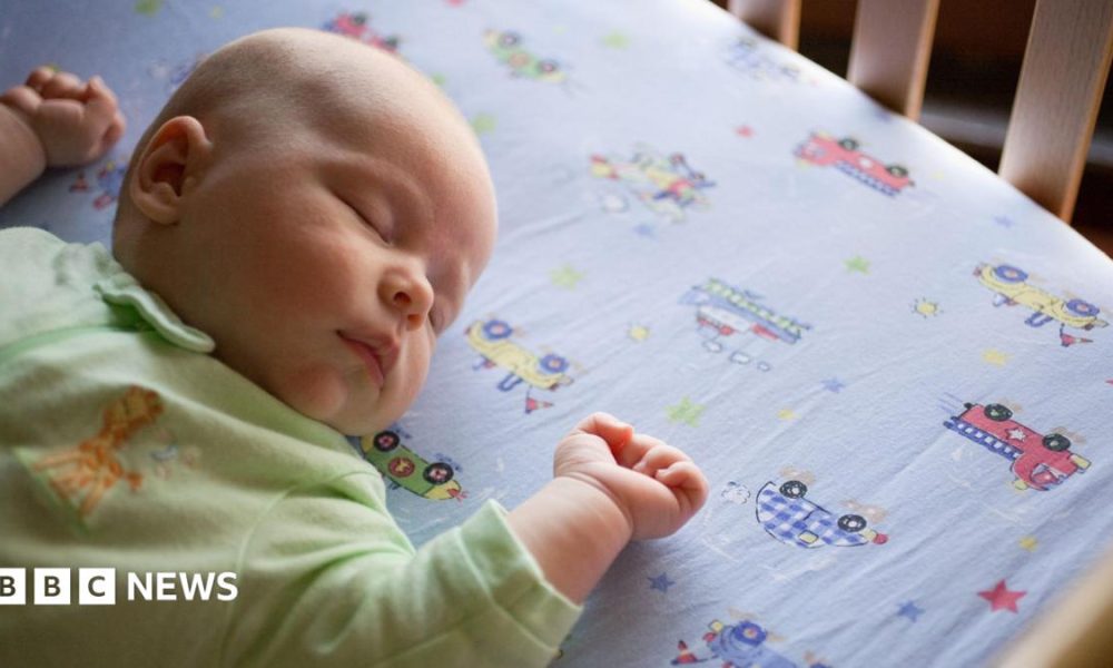 An overhead shot of a sleeping baby wearing a green onesie lying on a bedsheet with cars and trucks.