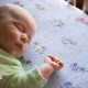 An overhead shot of a sleeping baby wearing a green onesie lying on a bedsheet with cars and trucks.
