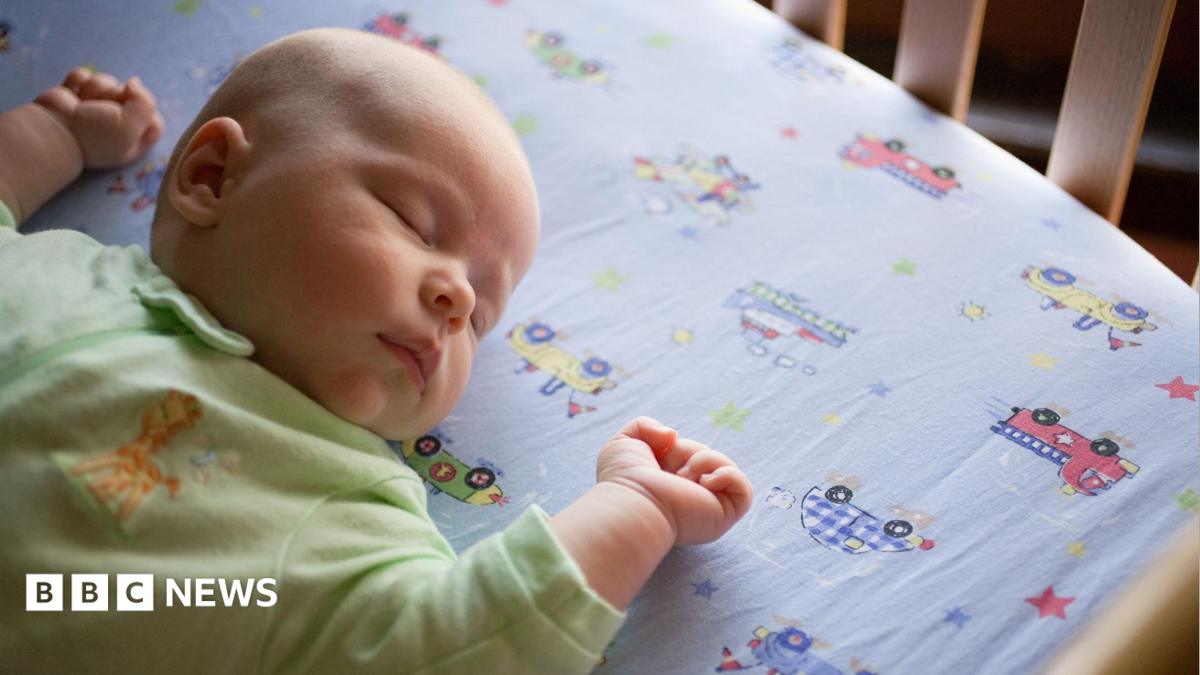 An overhead shot of a sleeping baby wearing a green onesie lying on a bedsheet with cars and trucks.