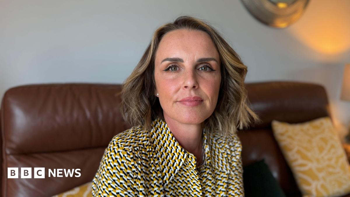 Rachel, who has blonde-brown hair, looks at the camera. She is sat on a brown leather sofa that is scattered with yellow and white cushions. She is wearing a black, white and yellow shirt.