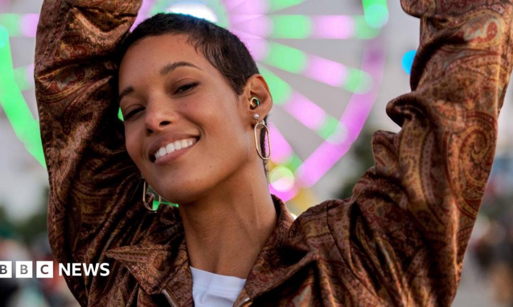 A young woman dances at dusk in front of a neon-lit Ferris wheel at a music festival. As well as large hoop earrings, she wears a rainbow-coloured ear-plug as protection for her hearing.