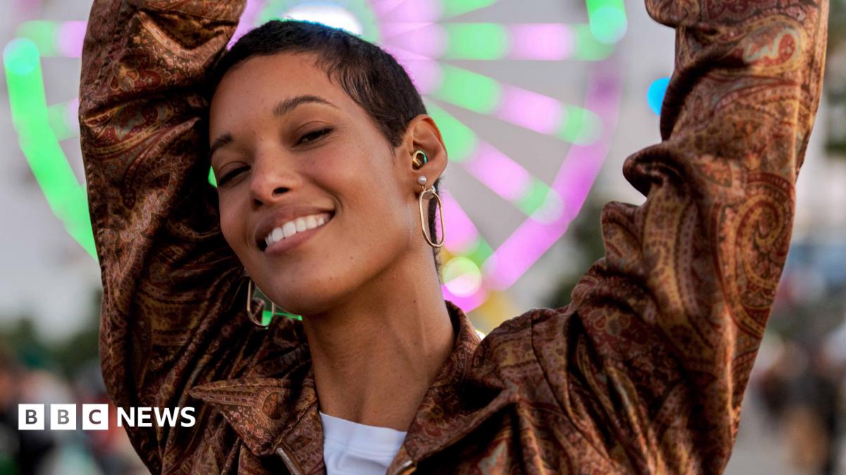 A young woman dances at dusk in front of a neon-lit Ferris wheel at a music festival. As well as large hoop earrings, she wears a rainbow-coloured ear-plug as protection for her hearing.
