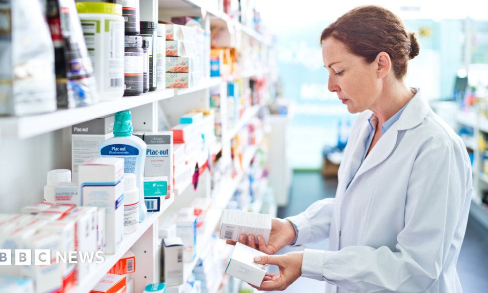 Pharmacist standing in front of shelves in pharmacy, holding medicine boxes in hands.