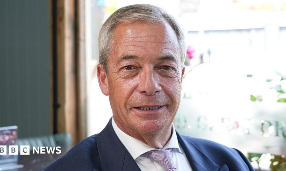 Nigel Farage sitting in a pub while wearing a white shirt and navy blazer. He is smiling.