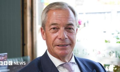 Nigel Farage sitting in a pub while wearing a white shirt and navy blazer. He is smiling.