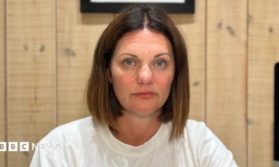 A woman with dark hair and blue eyes in a plain white T-shirt sits at a desk in a wood-panelled home office, facing the camera. A computer monitor, notebook, water bottle, phone and glasses are visible on the desk, with framed artwork hanging on the wall behind.